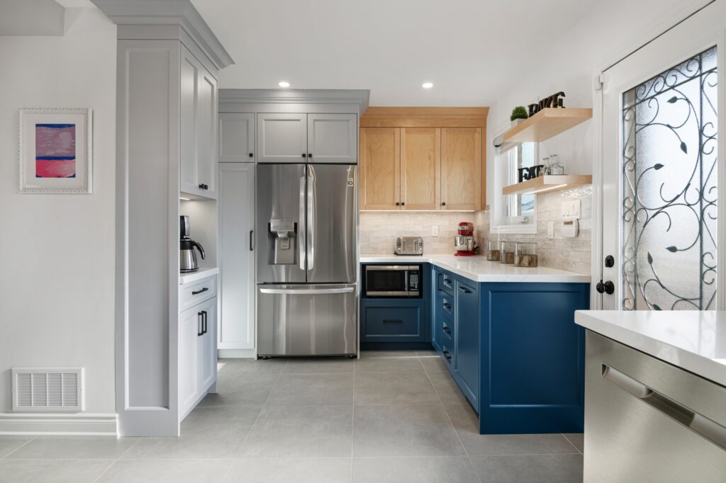 Modern kitchen with gray floor tiles, stainless steel appliances, light wood and blue cabinets, white countertops, and a decorative glass door. A coffee maker and mixer are visible on the counters.