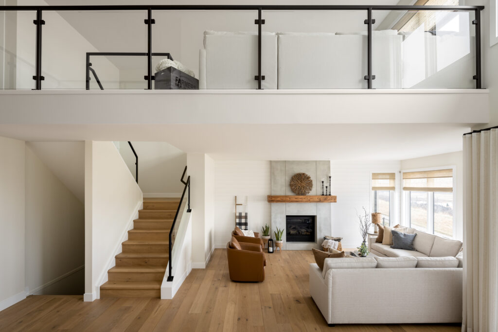 Modern, bright living room with light wood floors, white walls, a beige sectional sofa, two brown chairs, a fireplace with a wood mantel, large windows, and an open staircase leading to a loft with glass railing.