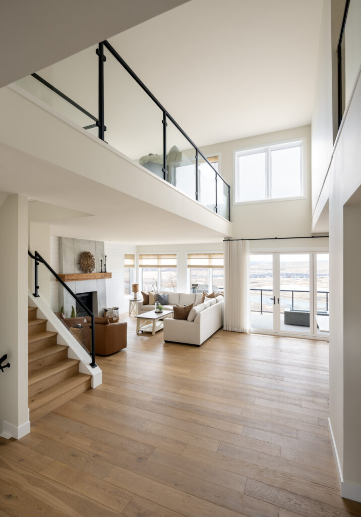 Spacious, modern living room with light wood floors, large windows, a fireplace, and neutral-colored furniture. An open loft and glass railing overlook the space, with stairs leading to the upper floor.