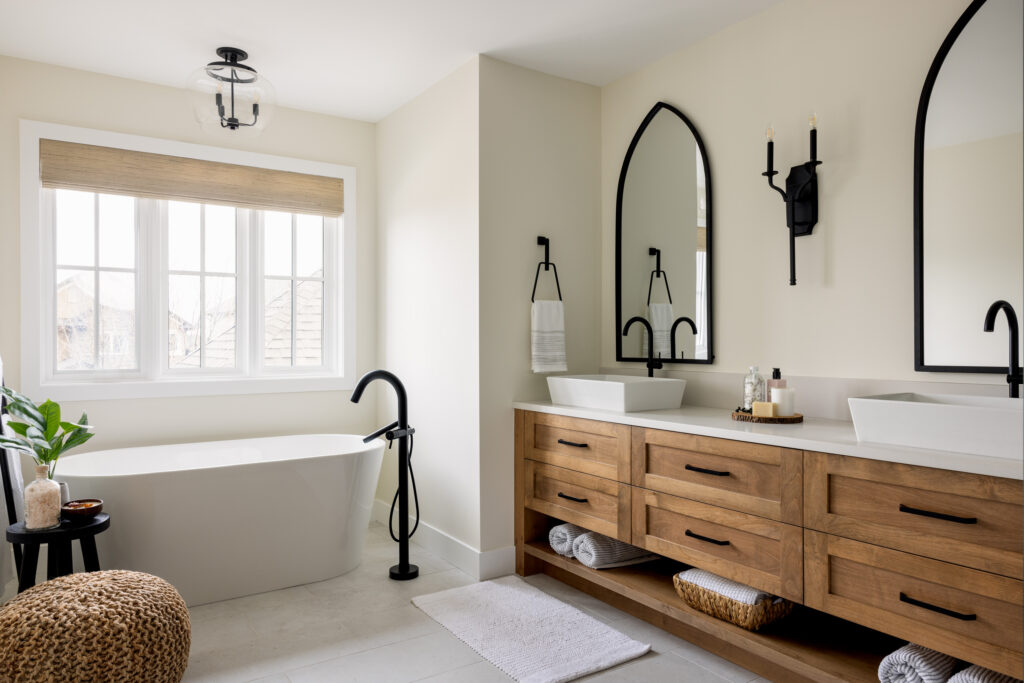 Modern bathroom with a freestanding tub, black fixtures, double sinks on a wooden vanity, large arched mirrors, wall sconces, a window with a woven shade, and minimalist decor.