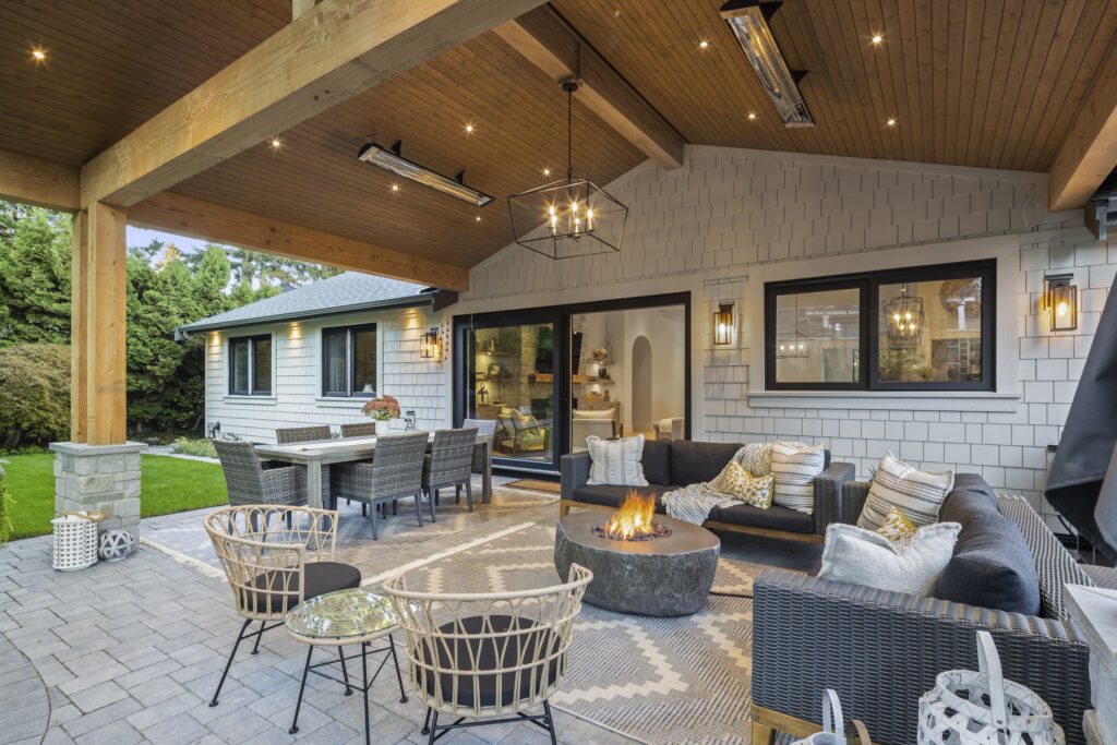 A covered patio with cozy seating, a fire pit, dining table, and modern decor; warm lighting and wooden beams create an inviting outdoor living space next to a house with gray siding.