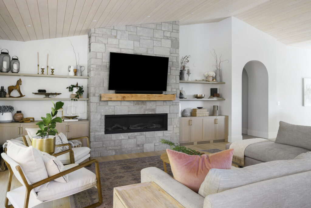 A modern living room with a stone fireplace, mounted TV, light wood furniture, shelves with decor, a gray sectional sofa, and potted plants; the space is bright with neutral tones and natural light.