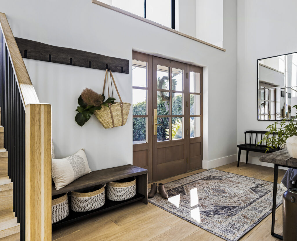 Bright, modern entryway with wooden double doors, a patterned rug, a black bench with pillows and baskets underneath, a wall hook holding a bag and foliage, and a staircase with wooden steps and black railing.