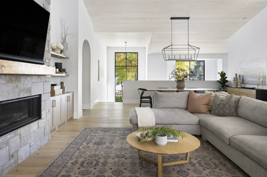Modern living room with a gray sectional sofa, wooden coffee table, patterned rug, stone fireplace, and wall-mounted TV. Large windows bring in natural light, and dining area is visible in the background.