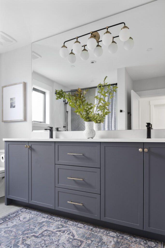Modern bathroom with navy blue vanity, brass hardware, large mirror, white countertop, vase with green branches, and a decorative light fixture above. A patterned rug lies on the floor and there’s framed art on the wall.