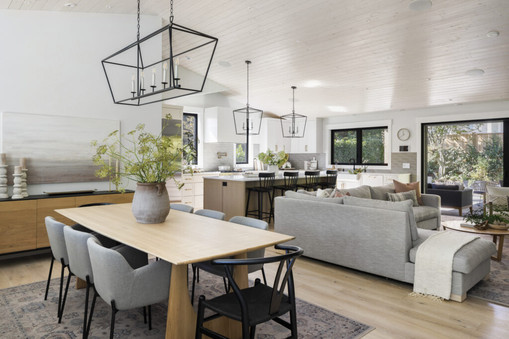 Open-concept living space with a wooden dining table and gray chairs, a modern kitchen with island seating, large windows, light wood floors, and cozy gray sofas arranged around a coffee table.