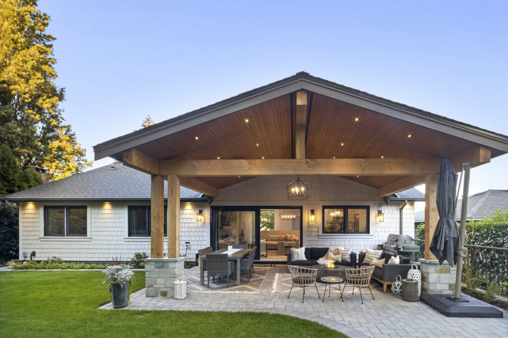 A modern house with a covered patio features outdoor seating, a fire pit, a dining table, and string lights. The yard is landscaped with grass, potted plants, and paving stones under a clear blue sky.