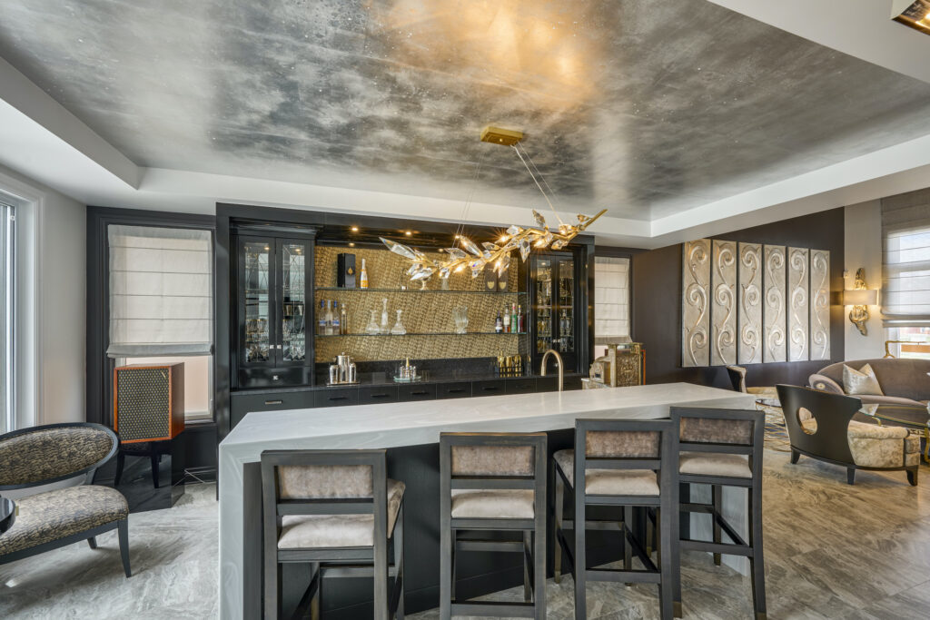 Modern home bar with a marble-topped island, four cushioned barstools, a black and gold backlit bar cabinet, contemporary chandelier, and elegant seating area in a luxurious, well-lit room.