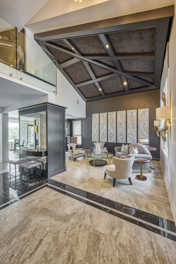 Spacious, modern living room with marble floors, high ceilings, stylish furniture, wall art panels, and glass balcony railing above. Large windows provide natural light, creating an elegant and airy atmosphere.