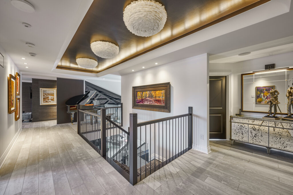 A modern upstairs hallway with wood floors, artwork on the walls, contemporary ceiling lights, a staircase with black railing, a mirrored console table, and a bronze sculpture. Natural light comes from a large window.