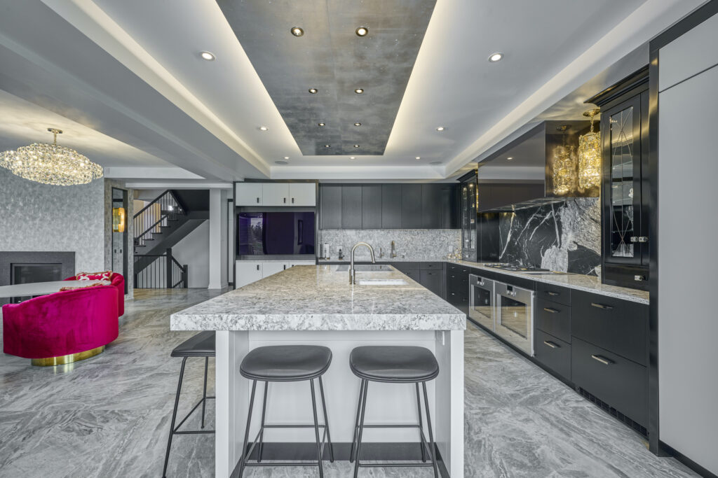 Modern kitchen with a large marble island, two stools, black cabinets, built-in appliances, a sleek ceiling with recessed lighting, and a glimpse of a red velvet chair and chandelier in the adjacent living area.