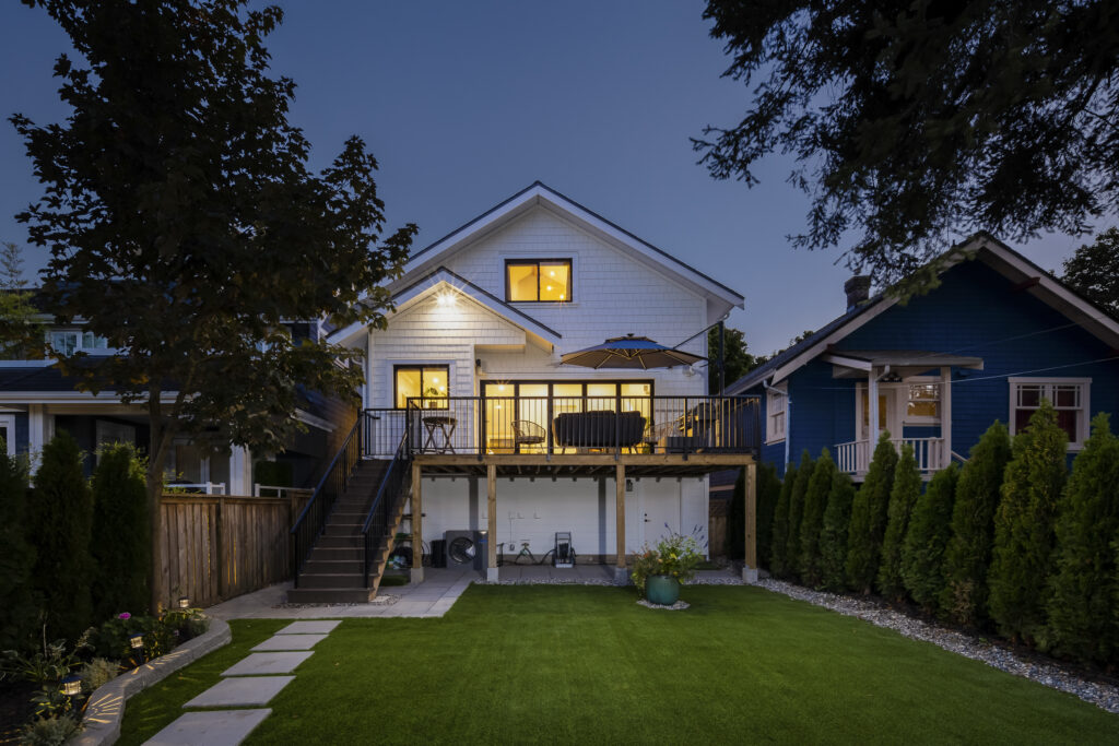 A two-story white house with a lit patio, balcony, and large windows, seen at dusk from a backyard with green grass, stepping stones, trees, and a neighboring blue house on the right.