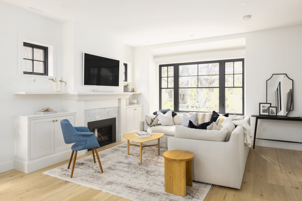 Bright, modern living room with a white sectional sofa, blue accent chair, small wooden tables, a fireplace, a wall-mounted TV, and large windows letting in natural light. A rug and decorative mirror add cozy touches.