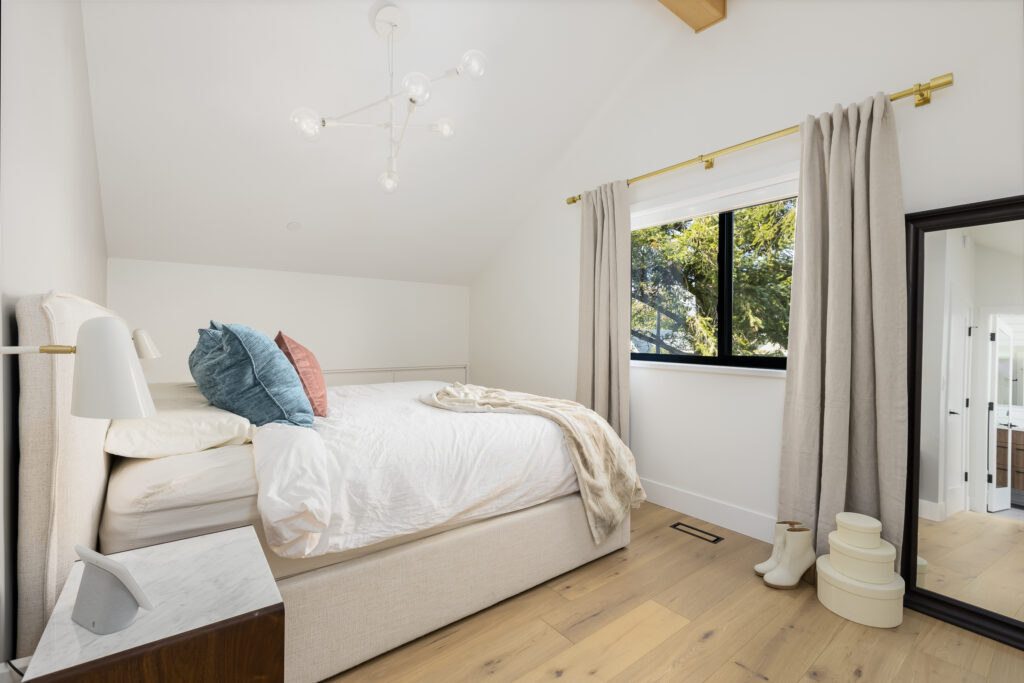 A modern, bright bedroom with a neatly made bed featuring white sheets and colorful pillows, a bedside table with a lamp, large window with beige curtains, a floor mirror, and wooden floors.