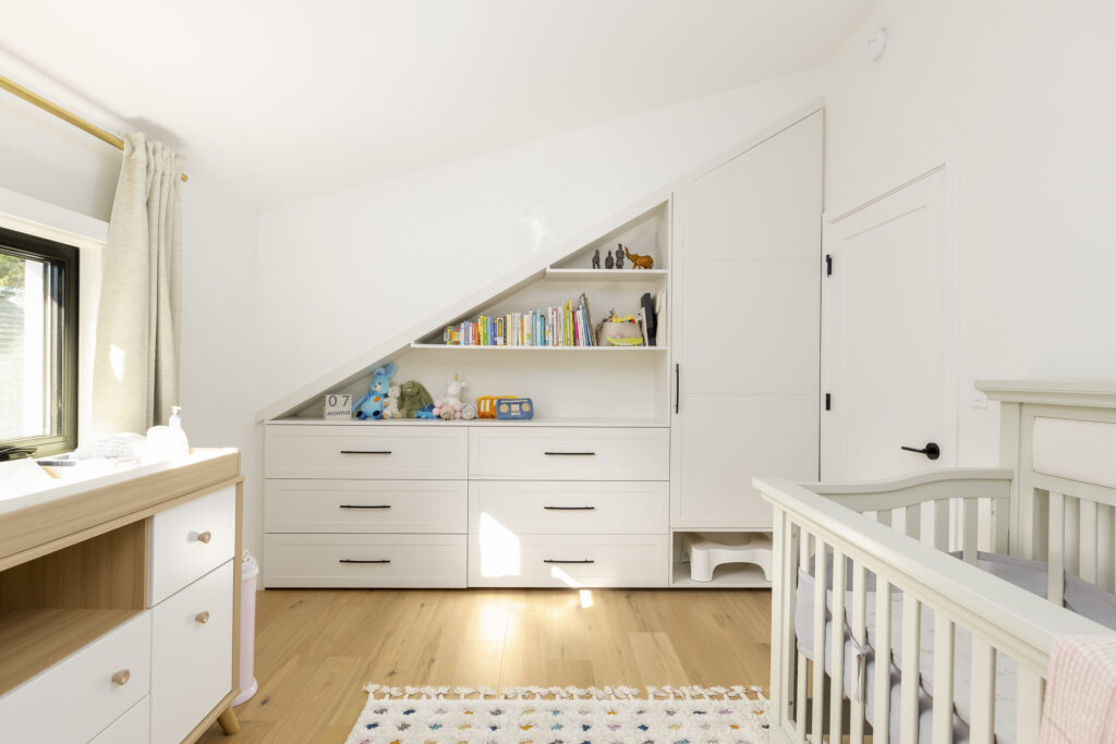 A bright, modern nursery with white furniture, a crib, dresser, and built-in shelves under a sloped ceiling, holding books, toys, and stuffed animals. Light wood floors and a window let in natural light.
