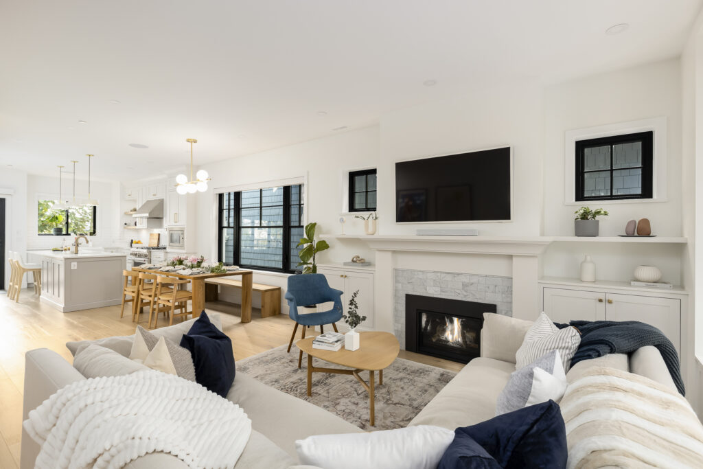 Modern open-concept living room and kitchen with light walls, wood floors, a white sectional sofa, blue accent chair, fireplace, wall-mounted TV, dining area, and large windows letting in natural light.