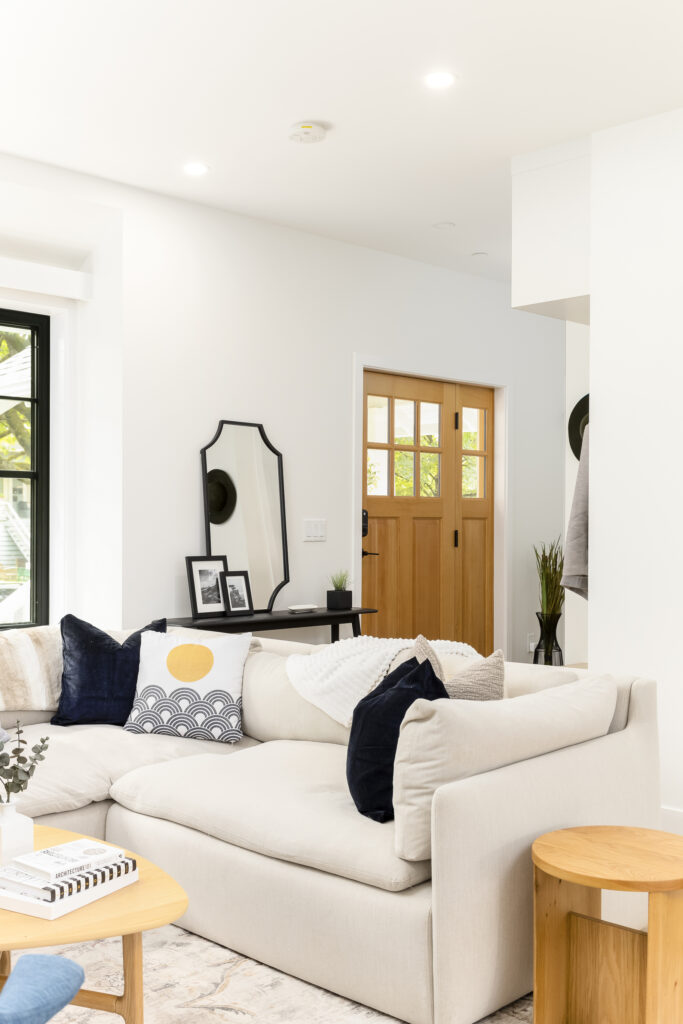 A bright, modern living room with a beige sectional sofa, dark and white throw pillows, a wooden side table, and decor including a mirror, framed photos, and plants. A wooden front door lets in natural light.