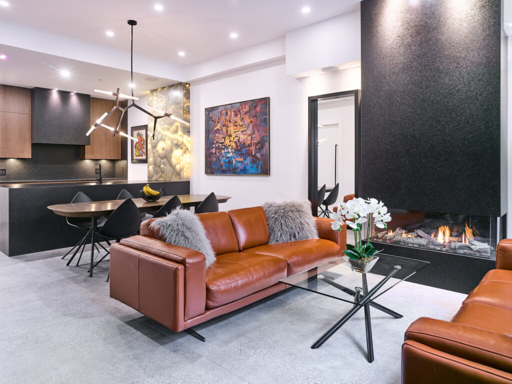 Modern living room with brown leather sofas, glass coffee table with white orchids, and a sleek black fireplace. In the background, a dining area with contemporary lighting and colorful abstract art on the walls.