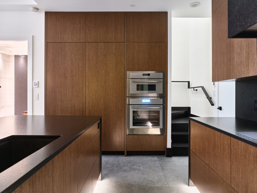 Modern kitchen with sleek wooden cabinets, black countertops, built-in stainless steel oven and microwave, and a staircase with black steps and railing in the background.