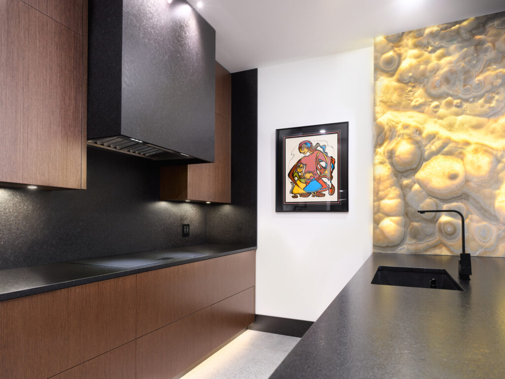 Modern kitchen with dark countertops, wood cabinets, a built-in stovetop, under-cabinet lighting, a black faucet, colorful framed artwork, and a textured, illuminated stone wall feature.