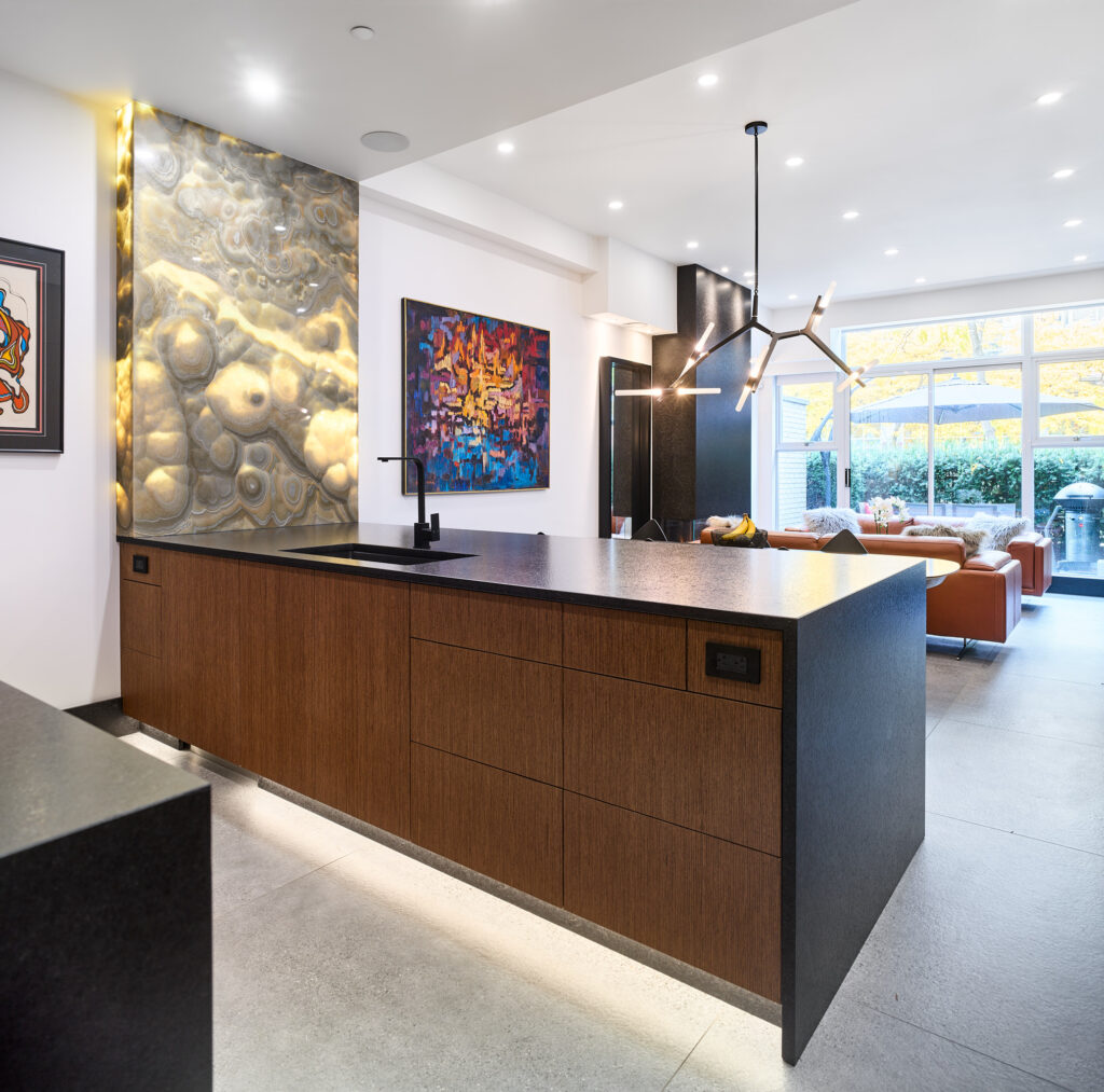 Modern kitchen with a large island featuring dark countertops and wood cabinets, abstract wall art, unique lighting fixtures, and an open living area with large windows and contemporary furniture.