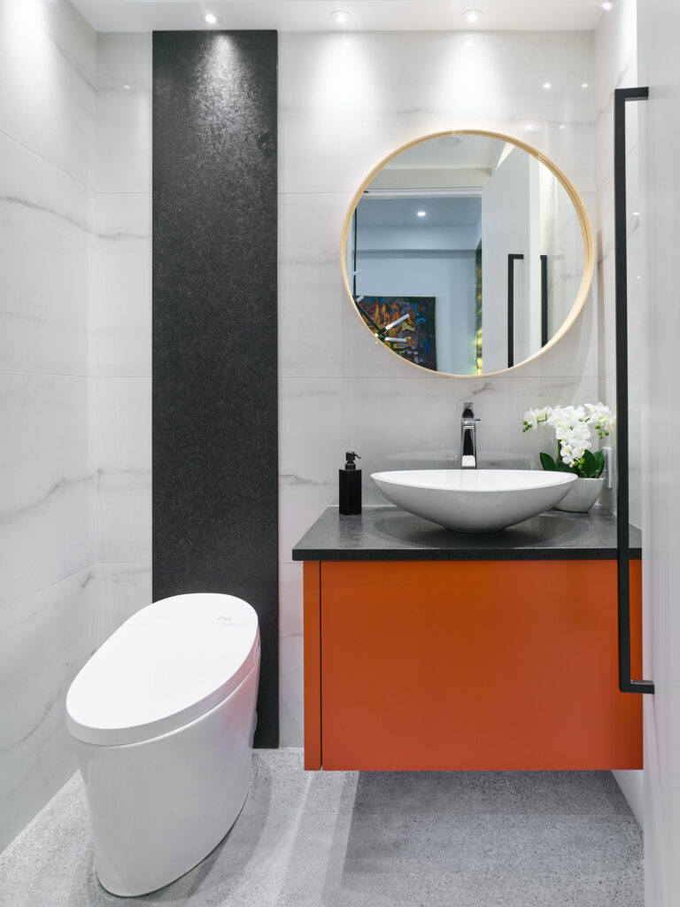 Modern bathroom with a white toilet, orange vanity, black countertop, vessel sink, round mirror, black soap dispenser, and white flowers; marble walls and recessed ceiling lights enhance the clean aesthetic.