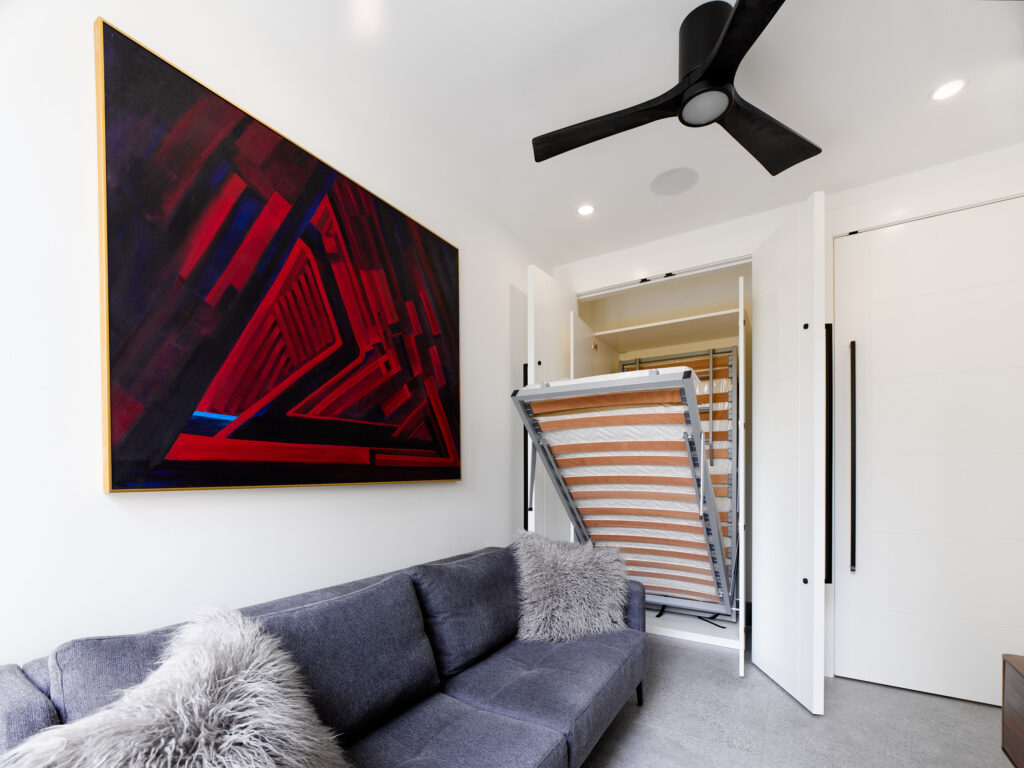 A modern room with a gray couch with fur pillows, a large abstract red and purple painting on the wall, a black ceiling fan, and a Murphy bed partially unfolded from a closet.