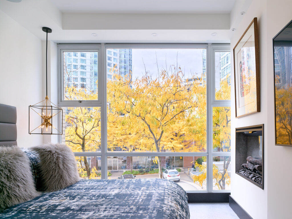 A modern bedroom with large windows showcasing trees with bright yellow autumn leaves and tall city buildings in the background. The room features a bed with textured pillows, a pendant light, a wall-mounted fireplace, and framed artwork.