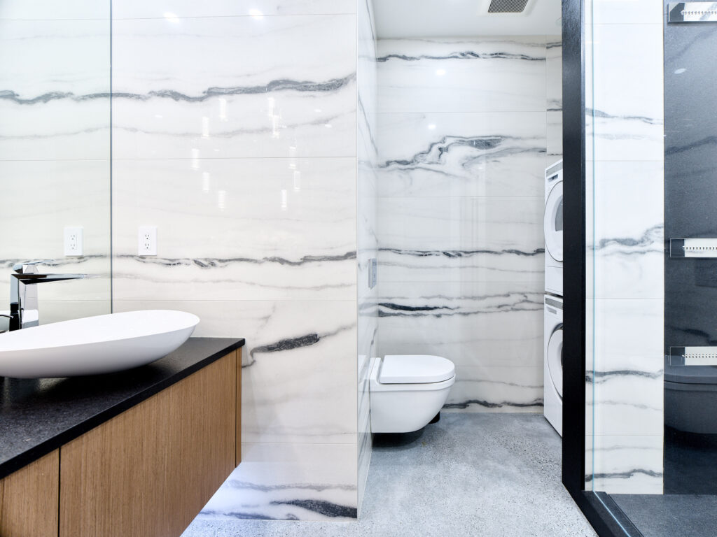 Modern bathroom with white and gray marble walls, floating wooden vanity with a vessel sink, wall-mounted toilet, stacked washer and dryer, and glass shower enclosure.