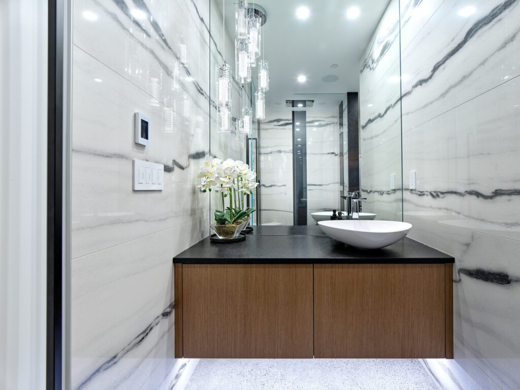 Modern bathroom with white marble walls featuring black veining, a floating wood vanity with a black countertop, a white vessel sink, a mirror, hanging glass light fixtures, and a potted orchid.