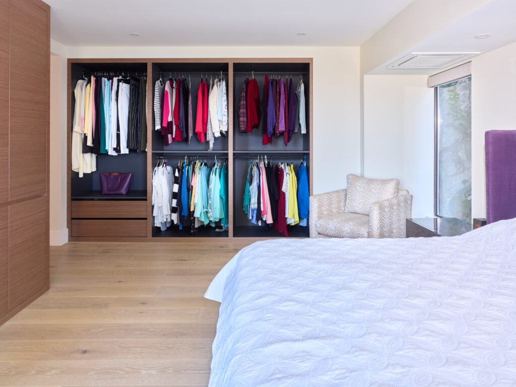 A modern bedroom with an open closet showing neatly hung clothes, a purple handbag, and wooden drawers. There is a bed with a white quilt, a patterned armchair, and a large window letting in natural light.