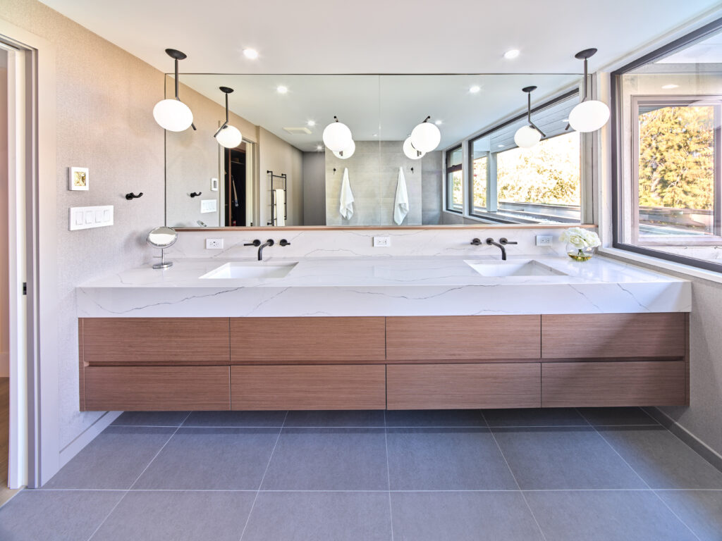 Modern bathroom with double sinks, a large mirror, hanging globe lights, wood vanity drawers, and large windows letting in natural light. The space features gray tile flooring and a clean, minimalist design.