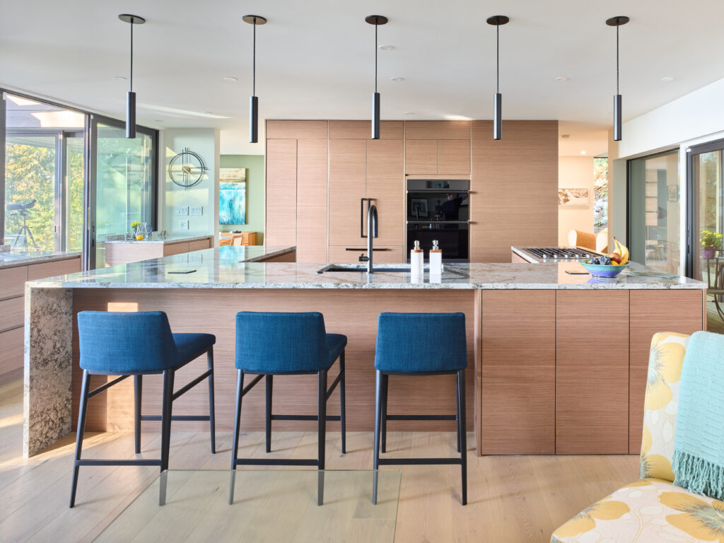 Modern kitchen with light wood cabinets, marble island countertop, three blue barstools, built-in oven, and black pendant lights. Large windows bring in natural light, and a cozy seating area is visible to the side.