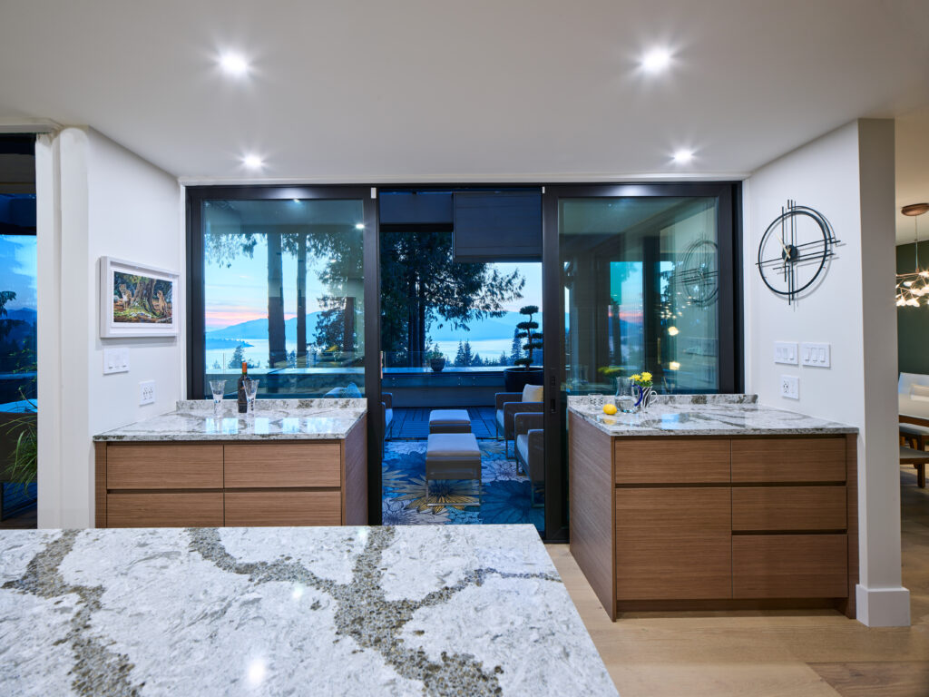 Modern kitchen with marble countertops, wooden cabinets, and large glass doors opening to a patio with outdoor seating. The view outside shows trees and a sunset over the water. Contemporary decor and bright lighting.