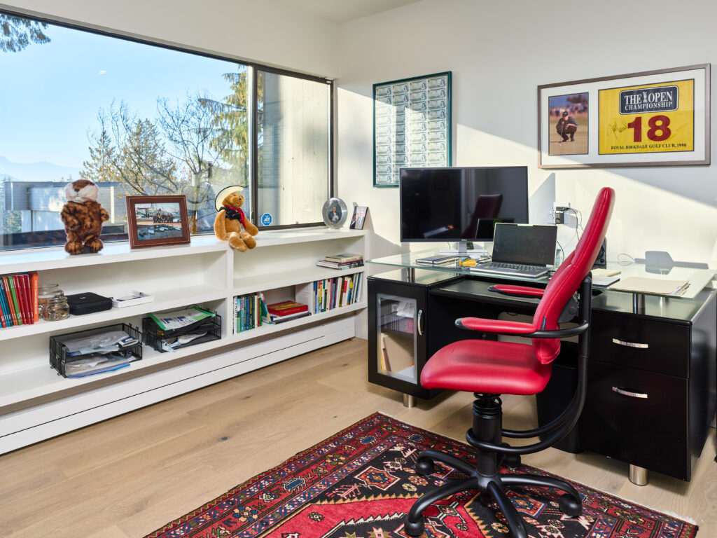 A modern home office with a black desk, red chair, computer monitors, and bookshelves. A window shows trees and mountains outside. The room has framed art, books, and stuffed animals on white shelves.