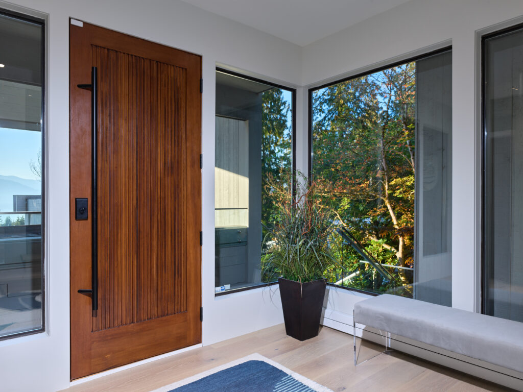 A modern entryway with a wooden front door, large windows letting in natural light, a potted plant, a gray bench, and light wood flooring. Trees and greenery are visible outside through the windows.