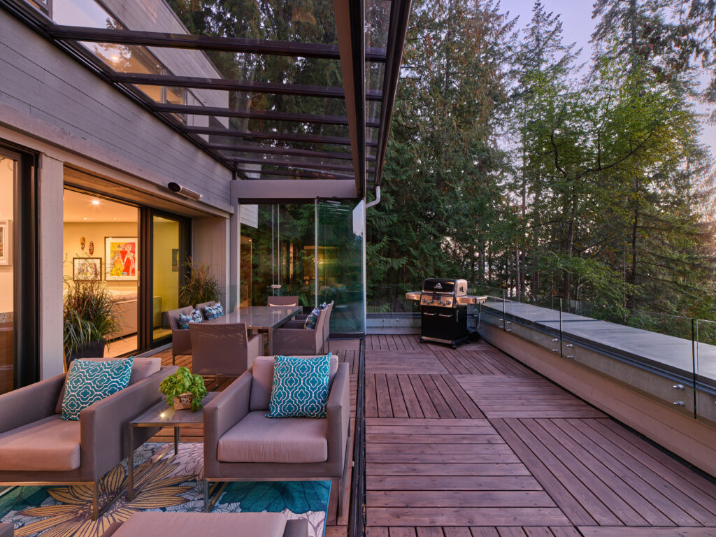 Modern outdoor patio with glass railing, wooden decking, lounge seating with blue patterned pillows, a barbecue grill, and surrounding trees, adjacent to a contemporary home with large windows and indoor art visible.