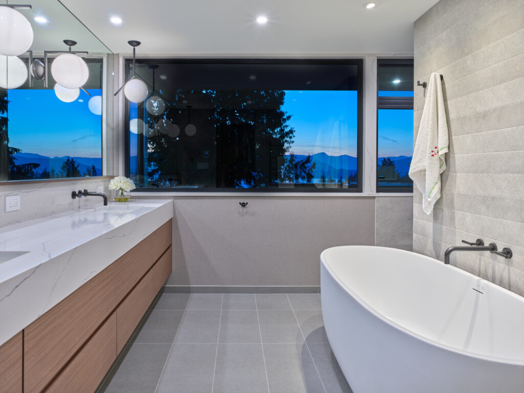 Modern bathroom with a freestanding white tub, wall-mounted faucet, large mirror, marble countertop, wooden cabinets, towel on a rack, and a large window showing a scenic evening view of trees and mountains.