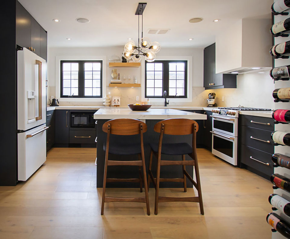 Modern kitchen with black cabinets, white countertops, wood floors, an island with two wooden chairs, a white fridge, a stove, large windows, pendant light, and a wall-mounted wine rack on the right.