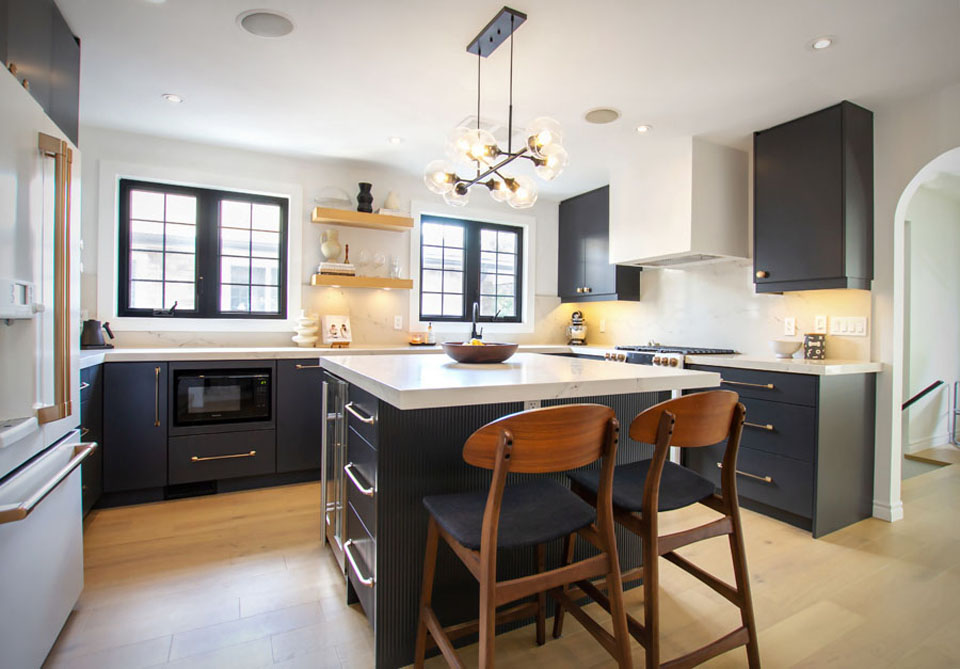 Modern kitchen with black cabinets, white countertops, a large island, wooden bar stools, light wood floors, and a contemporary chandelier. Bright natural light streams in from two large windows above the sink.