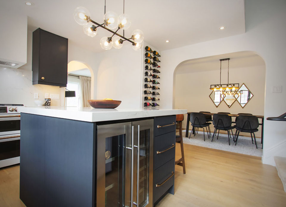 Modern kitchen with a navy island featuring a wine fridge and gold handles, a bowl on the counter, a wine rack on the wall, and a dining area with a geometric mirror and black chairs in the background.