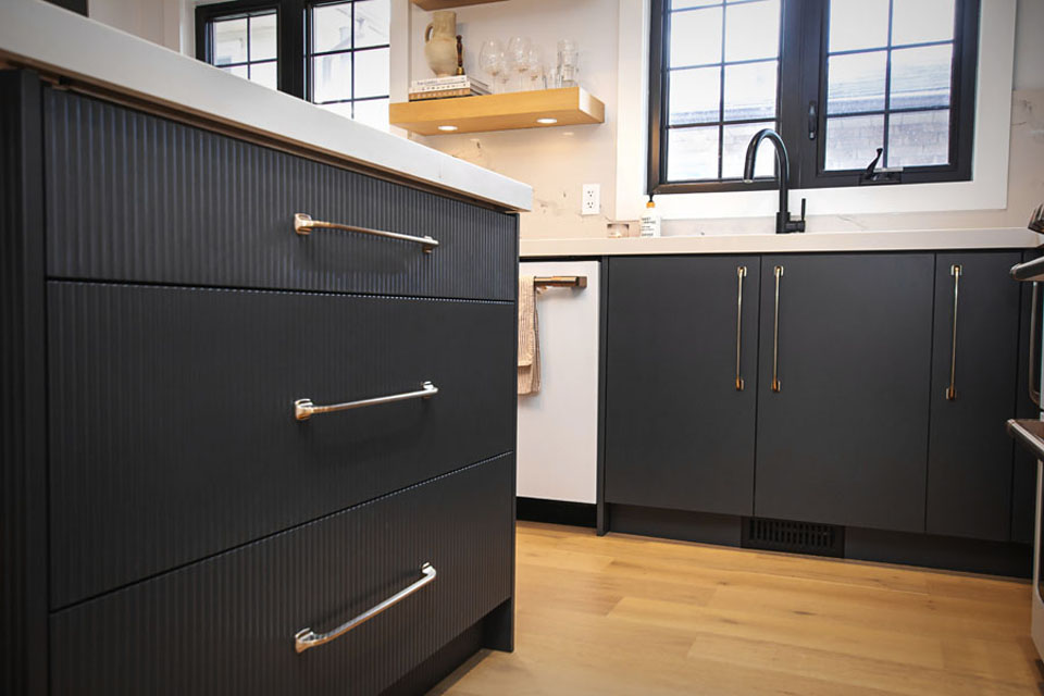 Modern kitchen with matte black cabinets, silver handles, a white countertop, black faucet, open shelves with glassware, and light wood flooring. Large windows provide natural light above the sink area.