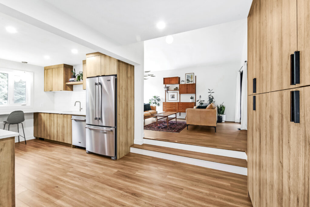 Modern kitchen with wood cabinets, stainless steel appliances, and white countertops, opening to a sunken living room with brown sofas, a wooden bookshelf, and indoor plants on hardwood floors. Natural light fills the space.