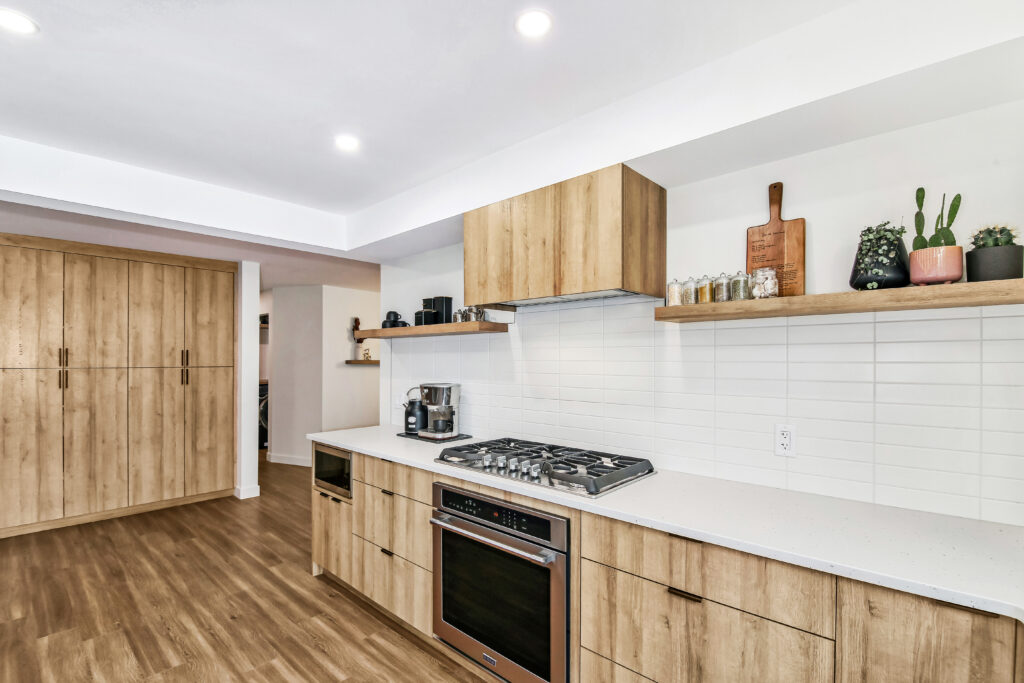 Modern kitchen with light wood cabinets, white countertops, built-in gas stove, oven, wall-mounted shelves holding jars and plants, white tile backsplash, and wood flooring under bright overhead lighting.