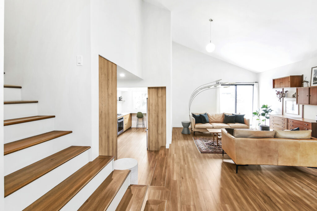 Modern living room with wooden floors, beige sofas, and a large arc floor lamp. An open kitchen is visible in the background, and wooden stairs lead upward on the left. The space is bright with natural light.