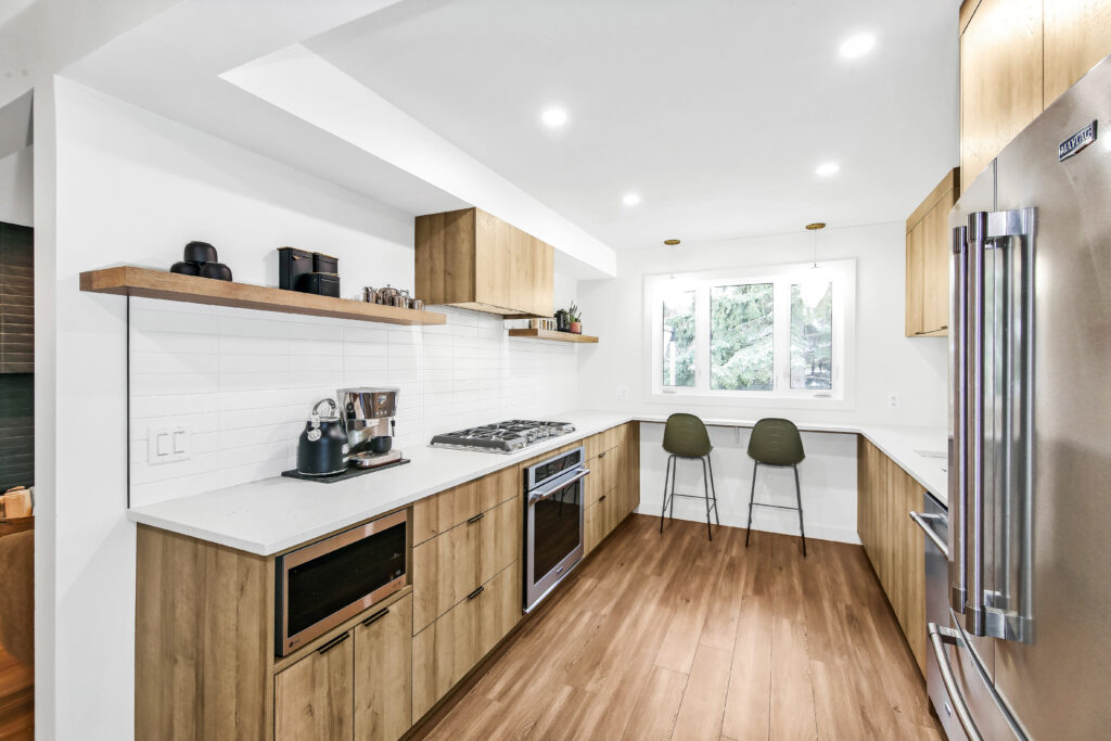 Modern kitchen with wooden cabinets, white countertops, built-in appliances, and a gas stove. Two green chairs are at a counter beneath a window, and a stainless steel refrigerator stands on the right. Bright, natural lighting.