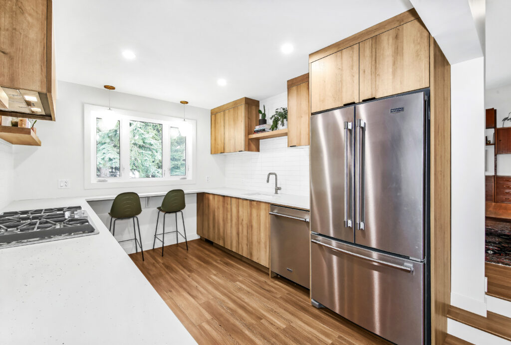 Modern kitchen with wood cabinets, stainless steel appliances, white countertops, a gas stove, and a double-door refrigerator. Two green chairs sit by a window with a view of trees outside. The floor is light wood.