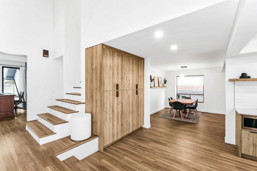 A modern, open-concept interior with light wood floors, a wooden staircase on the left, built-in wooden cabinets, and a dining area with a round table and chairs in the background. The space is bright and minimalist.