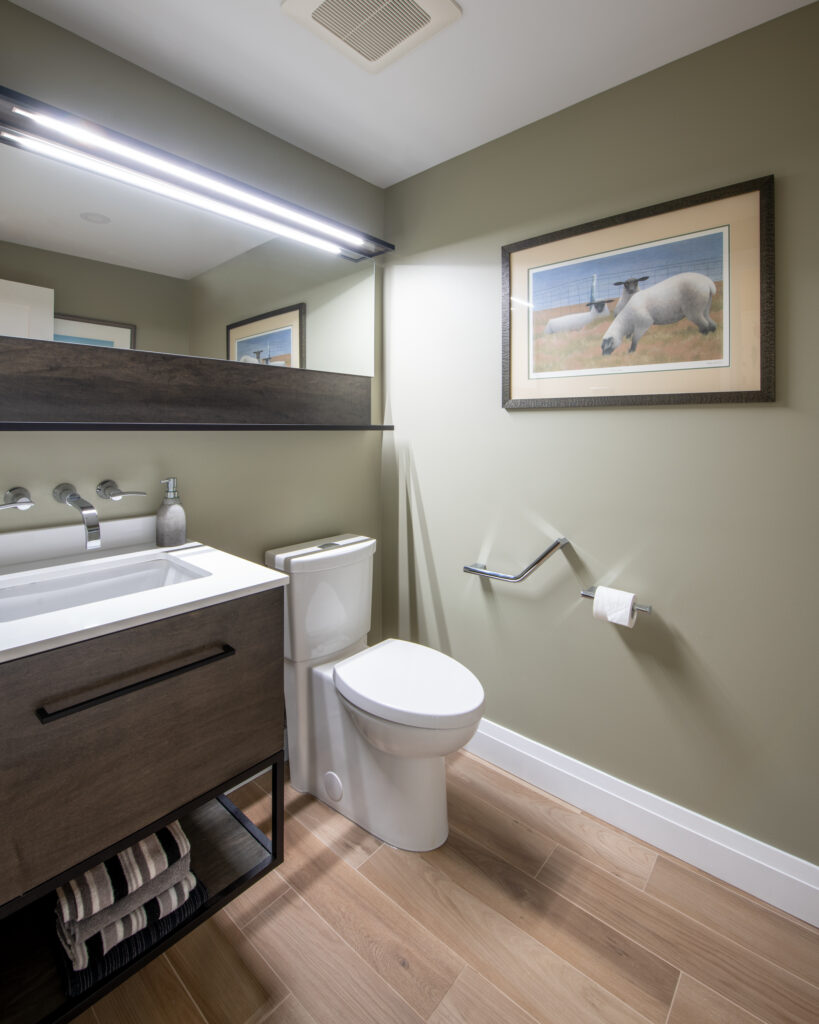 Modern bathroom with light wood floors, olive green walls, a white toilet, wall-mounted vanity with a mirror, towel shelf below, and a framed picture of a polar bear above the toilet. Toilet paper is mounted on the wall.