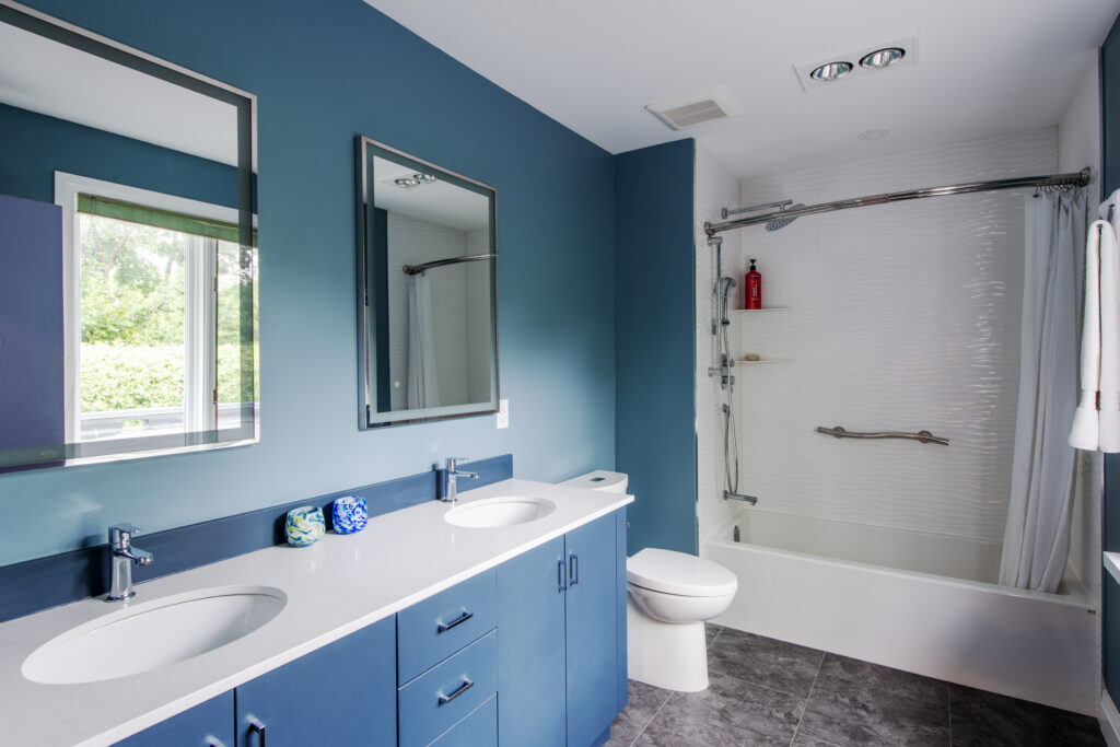Modern blue bathroom with double sinks, white countertops, large mirrors, a toilet, and a bathtub with a shower and white curtain. There is a window with greenery outside and gray tile flooring.